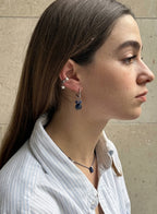 A close-up side profile of a woman with long brown hair wearing a pair of silver hoop earrings with dark blue stone drops, a silver star stud, and ear cuffs, paired with a light blue and white striped shirt.