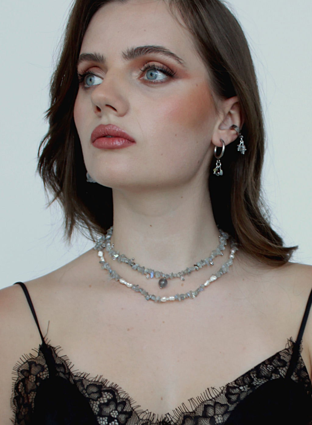 A close-up of a woman wearing a double-layered silver rhinestone choker necklace and matching silver hoop earrings with dangling clear crystal charms, styled with a black lace top against a plain light background.