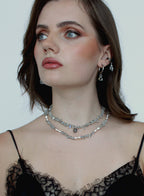 A close-up of a woman wearing a double-layered silver rhinestone choker necklace and matching silver hoop earrings with dangling clear crystal charms, styled with a black lace top against a plain light background.