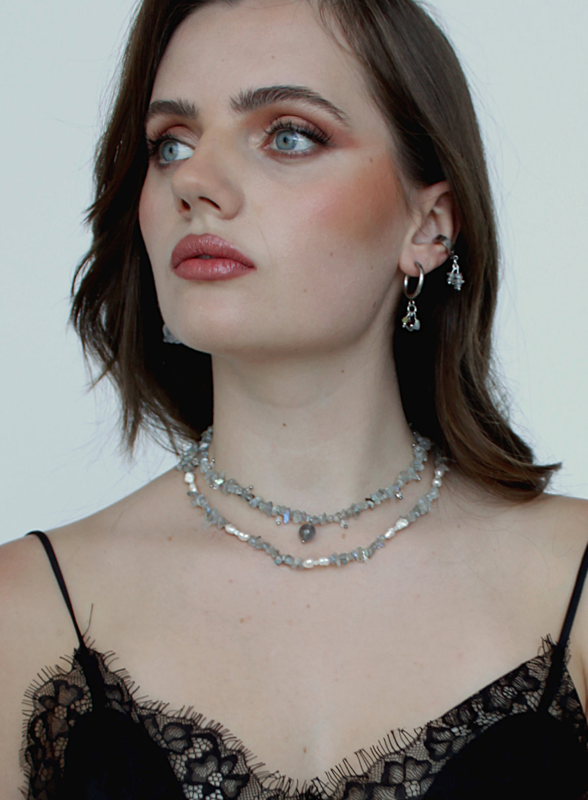 A close-up of a woman wearing a double-layered silver rhinestone choker necklace and matching silver hoop earrings with dangling clear crystal charms, styled with a black lace top against a plain light background.