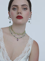 A close-up of a model wearing a bohemian-style layered necklace and matching drop earrings. The jewelry features light green beads, small dark accents, and a lavender purple pendant, styled with a white lace-trimmed top against a plain light gray background.
