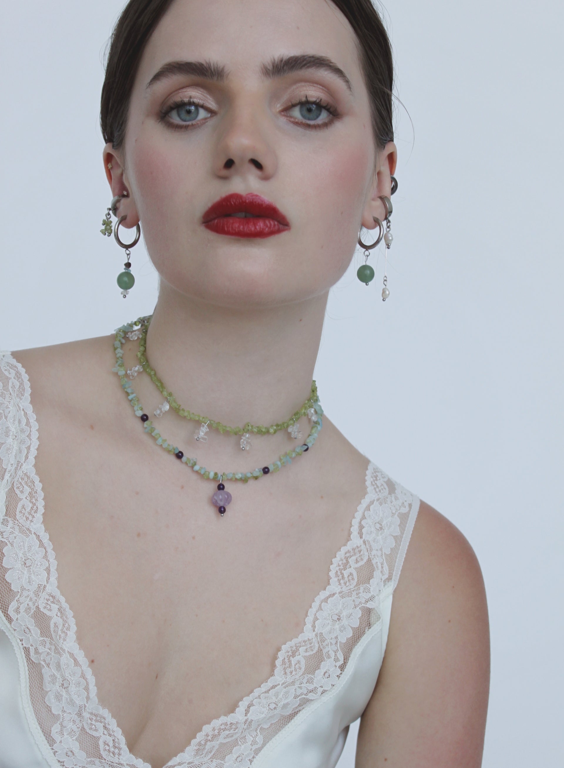 A close-up of a model wearing a bohemian-style layered necklace and matching drop earrings. The jewelry features light green beads, small dark accents, and a lavender purple pendant, styled with a white lace-trimmed top against a plain light gray background.