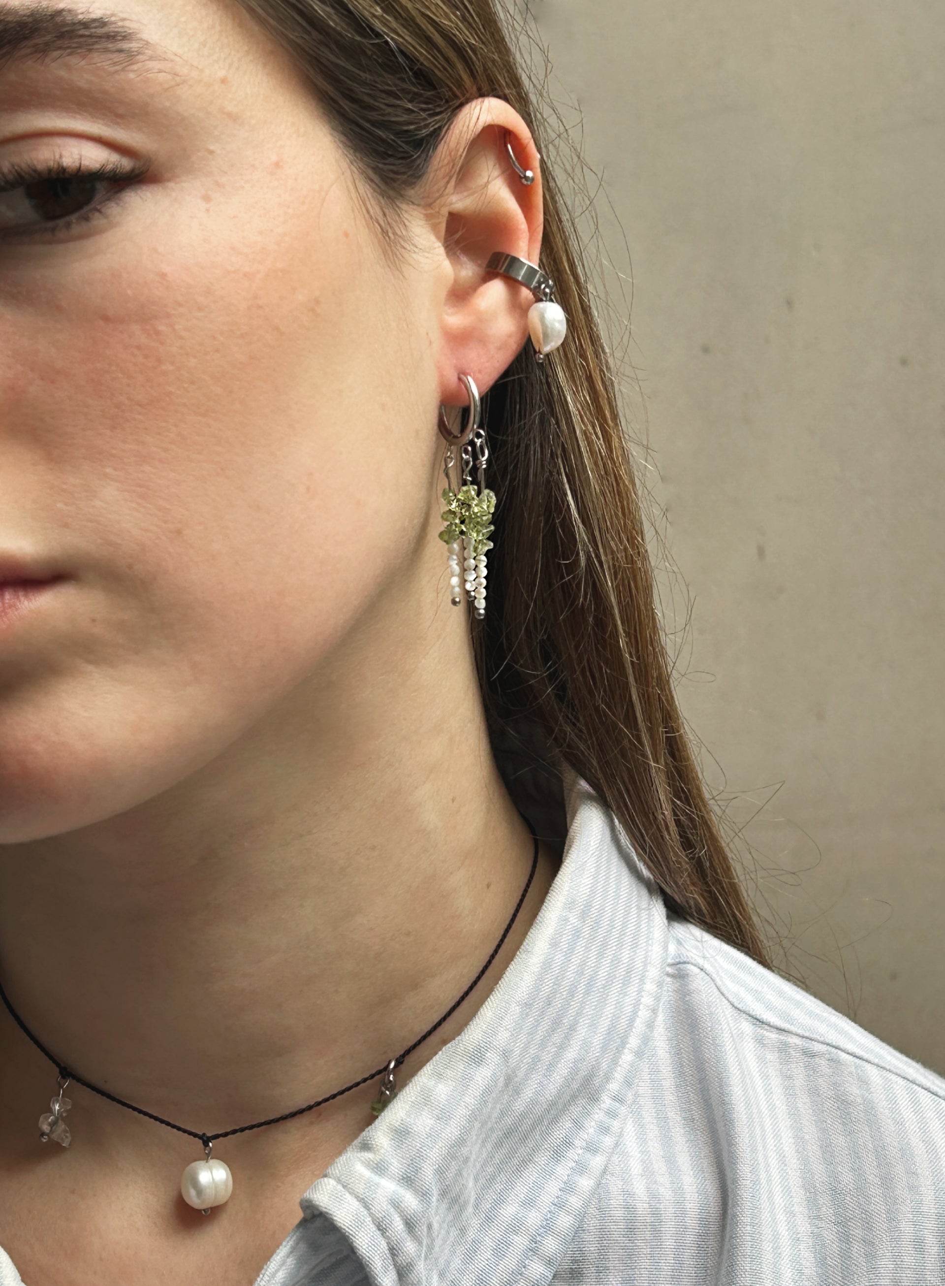 A close-up of a woman's ear wearing a silver hoop earring with a dangling green floral charm and clear crystal accents, alongside other ear piercings.