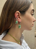A close-up side profile of a woman wearing silver hoop earrings with dangling green crystal-like charms, along with a matching green beaded necklace, against a neutral beige wall.
