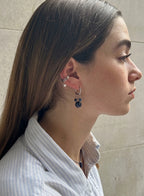A close-up side profile of a woman wearing a silver hoop earring with a dangling dark blue beaded mouse-head charm, along with other ear piercings, set against a neutral tiled wall.