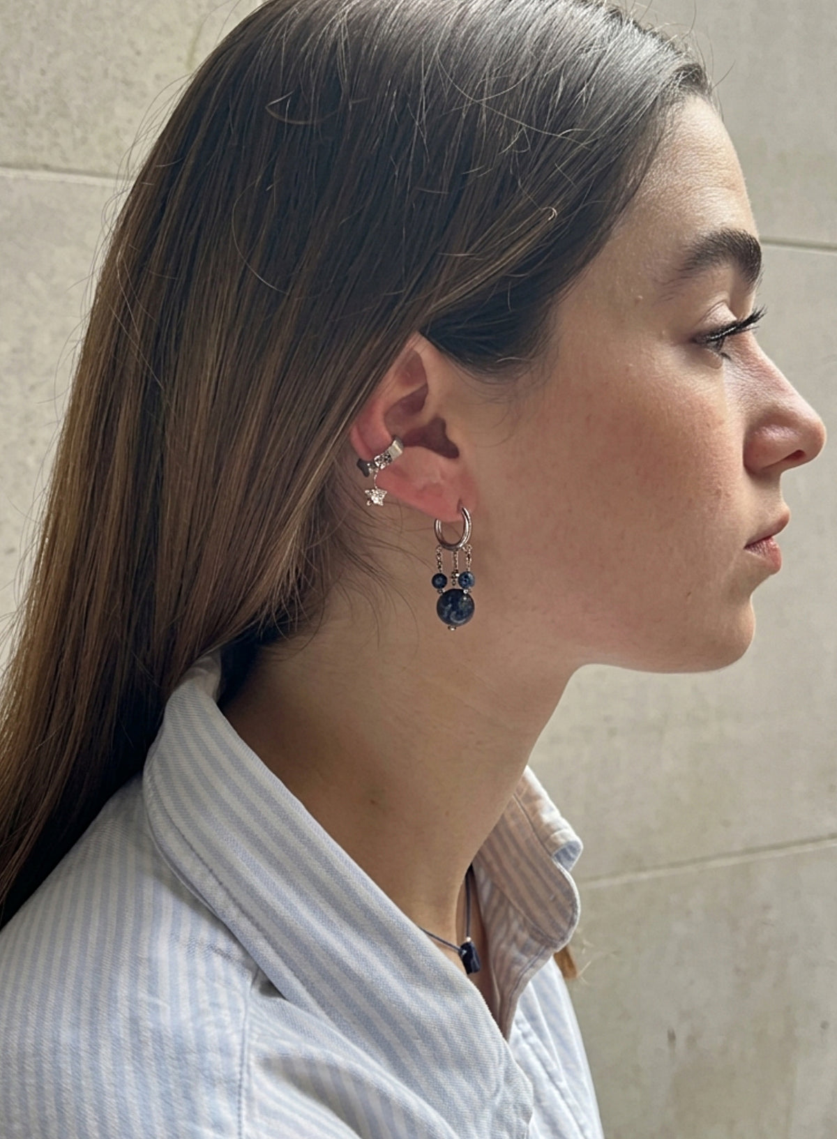 A close-up side profile of a woman wearing a silver hoop earring with a dangling dark blue beaded mouse-head charm, along with other ear piercings, set against a neutral tiled wall.