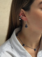 A close-up of a person's ear wearing a silver hoop earring with a small dangling charm and a silver hook earring with a dark blue faceted bead drop. The person is wearing a light blue and white striped shirt.