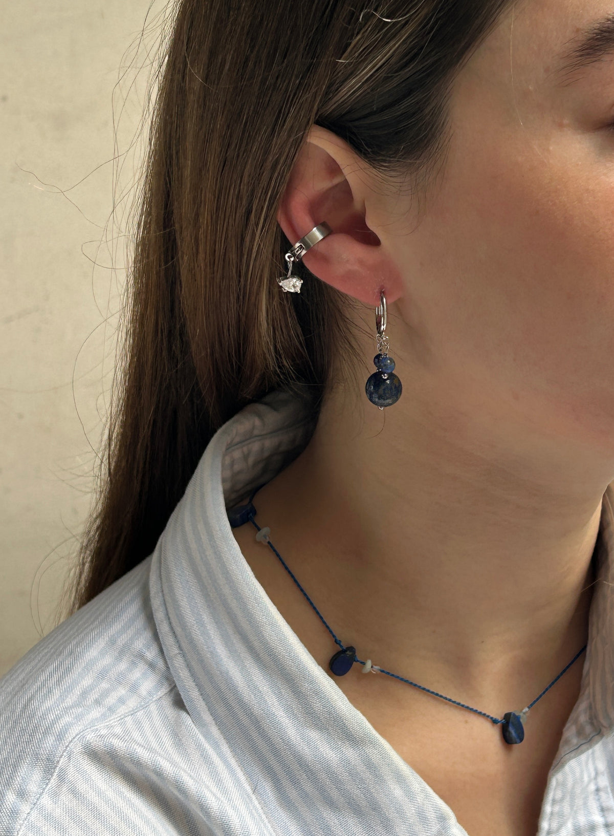 A close-up of a person's ear wearing a silver hoop earring with a small dangling charm and a silver hook earring with a dark blue faceted bead drop. The person is wearing a light blue and white striped shirt.