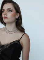 A woman with brown hair and blue eyes wearing silver beaded layered necklaces and matching drop earrings, paired with a black lace-trim camisole top, set against a plain light gray background.