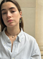 A close-up portrait of a woman wearing dangling earrings with green and white beads, a black cord necklace with a pearl, and a light blue striped button-up shirt.