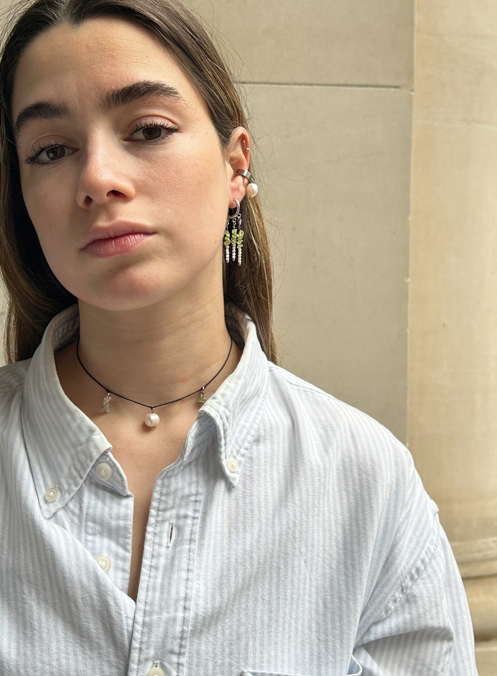 A close-up portrait of a woman wearing dangling earrings with green and white beads, a black cord necklace with a pearl, and a light blue striped button-up shirt.