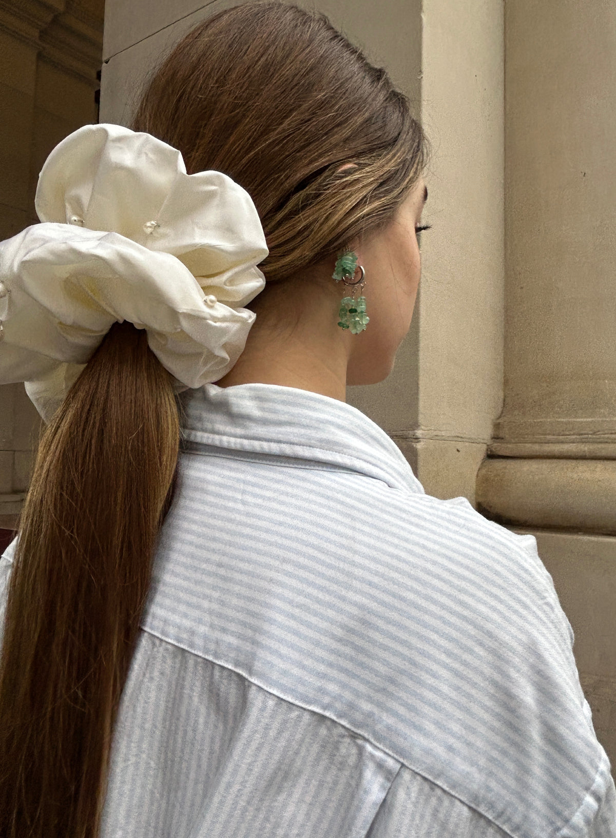 A woman shown from the back wearing a very large cream silk  scrunchie in her long brown hair, paired with a light blue and white striped shirt and green earrings, set against a beige stone architectural background.
