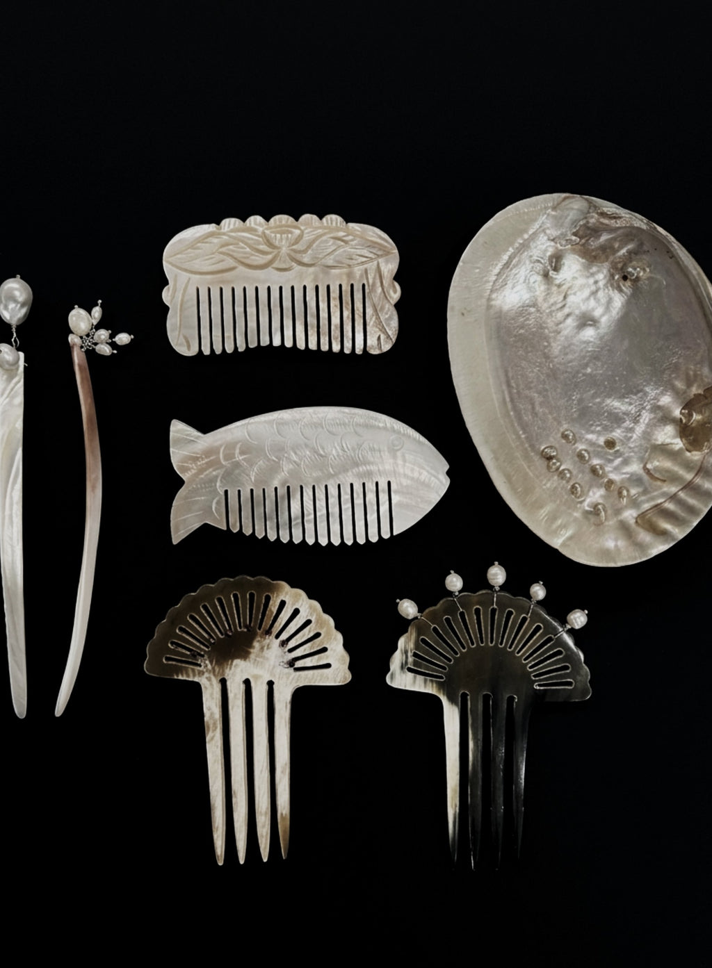 A collection of vintage hair accessories and combs displayed against a solid black background, including carved ivory or bone combs, pearl embellishments, and a large mother-of-pearl dish.