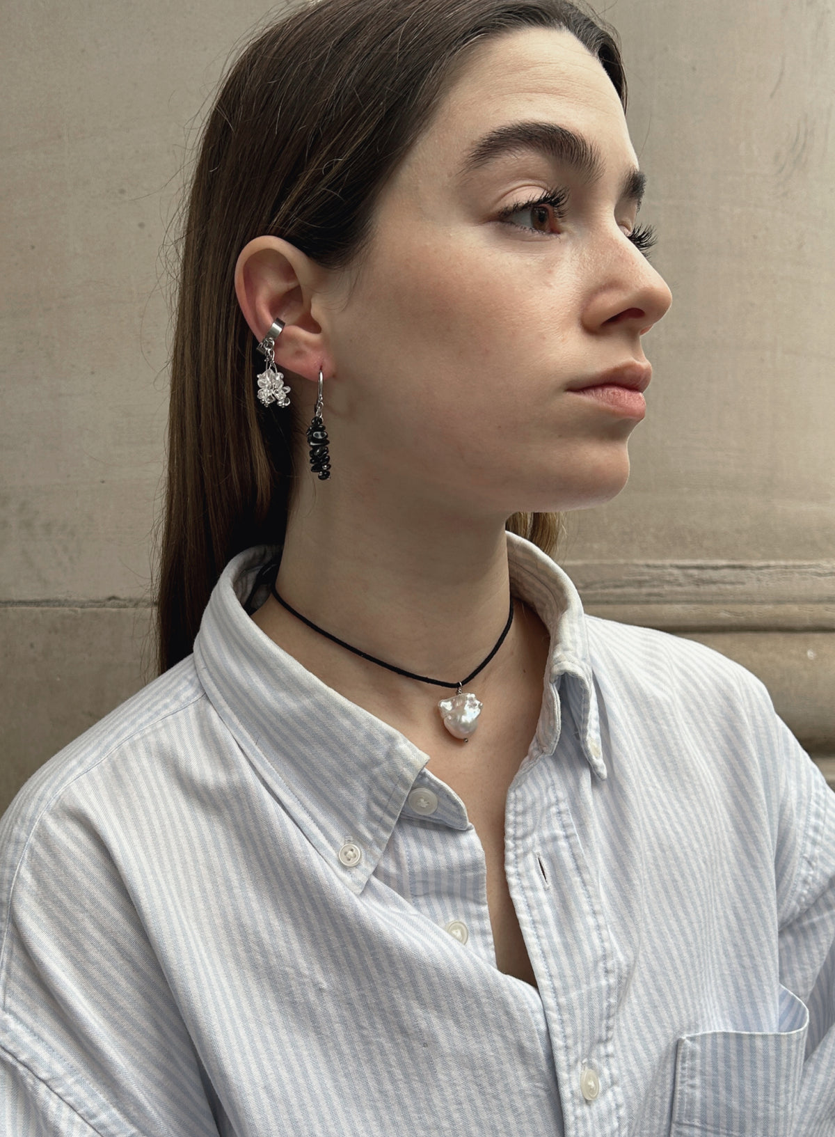 A woman in a light blue and white striped button-down shirt is shown in profile, wearing a black cord necklace with a white heart pendant and matching dangling earrings.