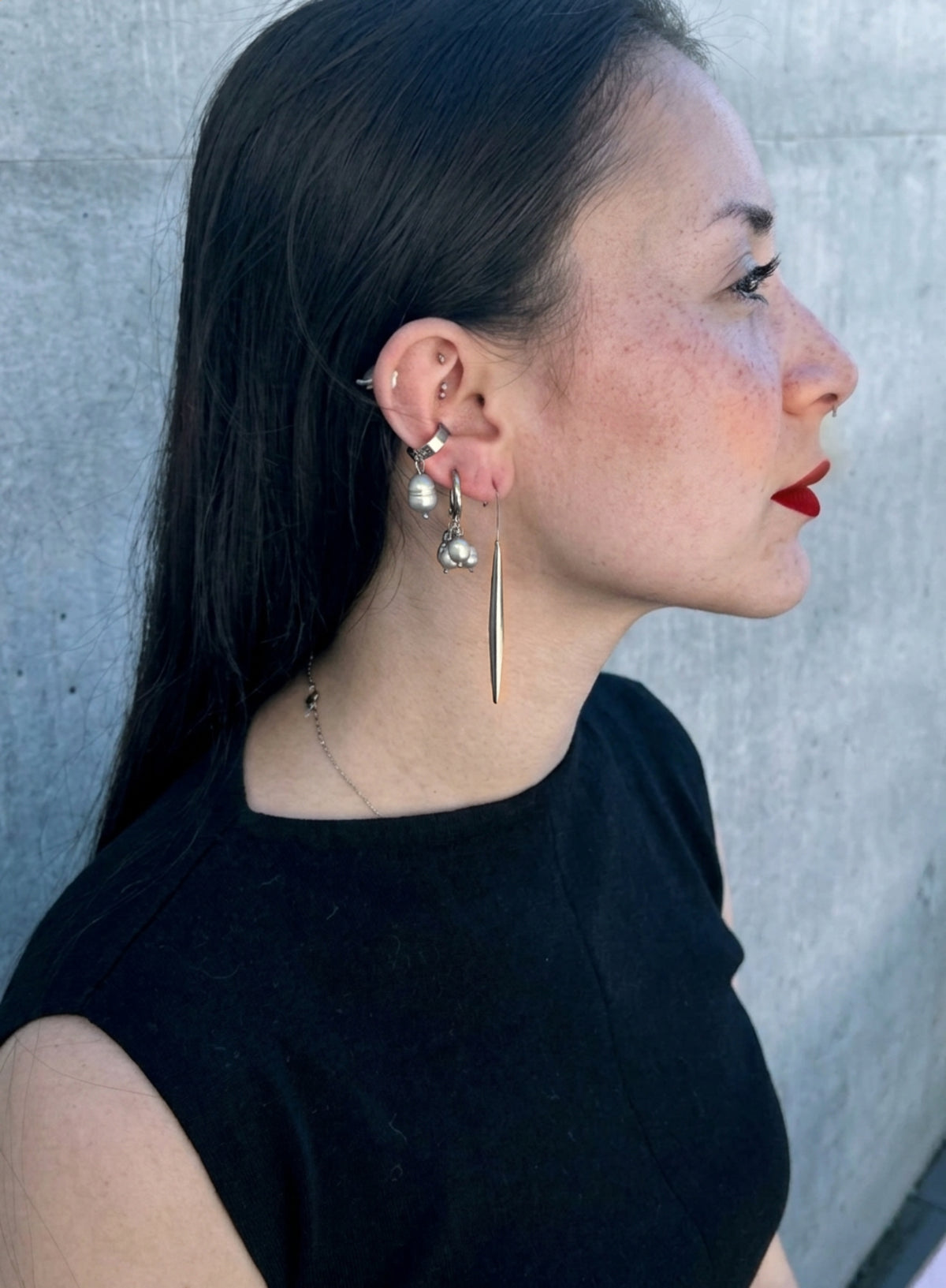 a woman with red lips and black dress wearing a pearl of grey pearl charms earrings in huggie hoop, long structural shape earring, and a large grey pearl ear cuff against concrete background