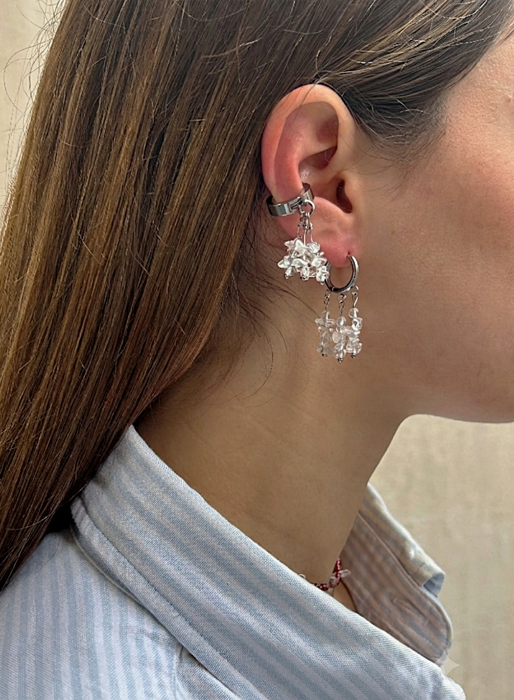 A close-up side profile of a woman wearing a silver-toned ear cuff with dangling clear crystal snowflake charms and a small hoop earring.