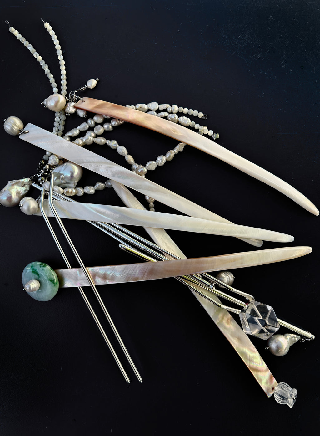 A set of traditional hair sticks made of mother-of-pearl, featuring iridescent white, beige, and green curved pieces, silver wire hairpins, and pearl bead strands, arranged on a solid black background.