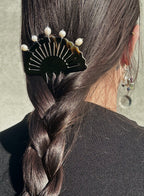 A close-up lifestyle shot of a model's dark brown hair styled in a side braid, secured with a black fan-shaped hair claw clip adorned with white pearl beads. The model is wearing a black ribbed top and a dangling beaded earring against a neutral concrete background.