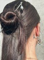 A close-up of a person's head from the back, showcasing a dark brown half-up bun styled with silver hairpins, one of which features a white pearl. The person also wears silver hoop earrings with dangling charms.