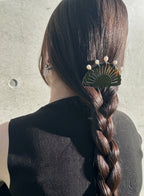 A woman viewed from behind wearing a long dark brown braided ponytail hair accessory with a fan-shaped gold and green hair clip adorned with small white pearl-like beads, set against a textured light gray wall.