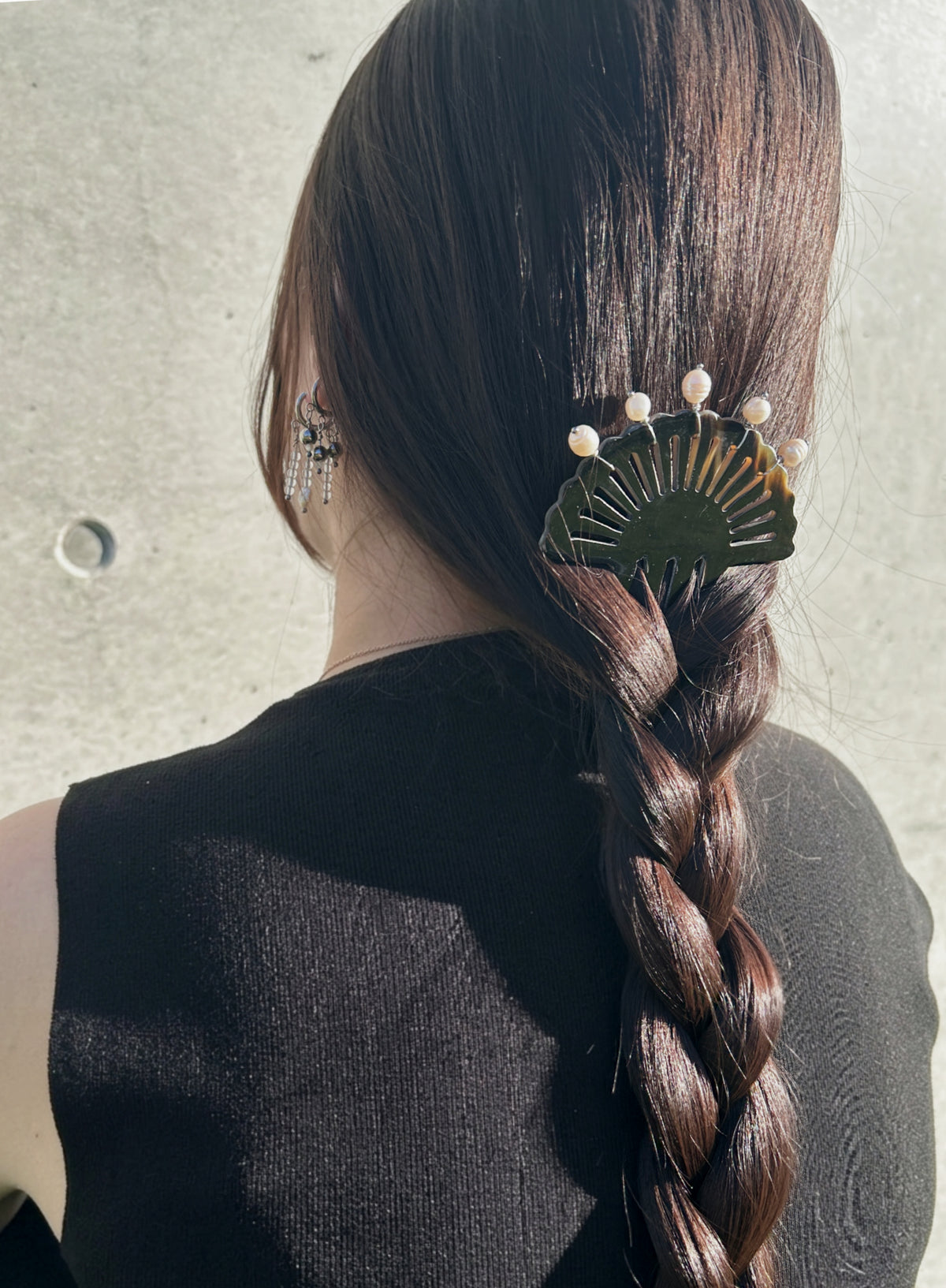 A woman viewed from behind wearing a long dark brown braided ponytail hair accessory with a fan-shaped gold and green hair clip adorned with small white pearl-like beads, set against a textured light gray wall.