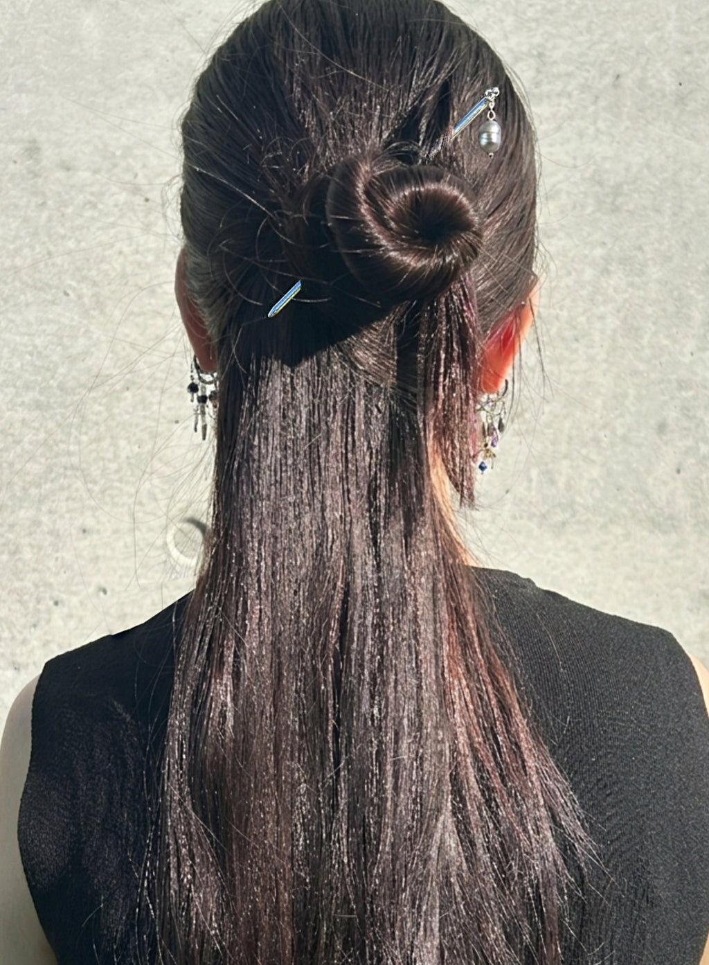 A person shown from the back with long, straight dark brown hair styled in a low bun with decorative hairpins and dangling earrings, wearing a black top against a light gray wall.
