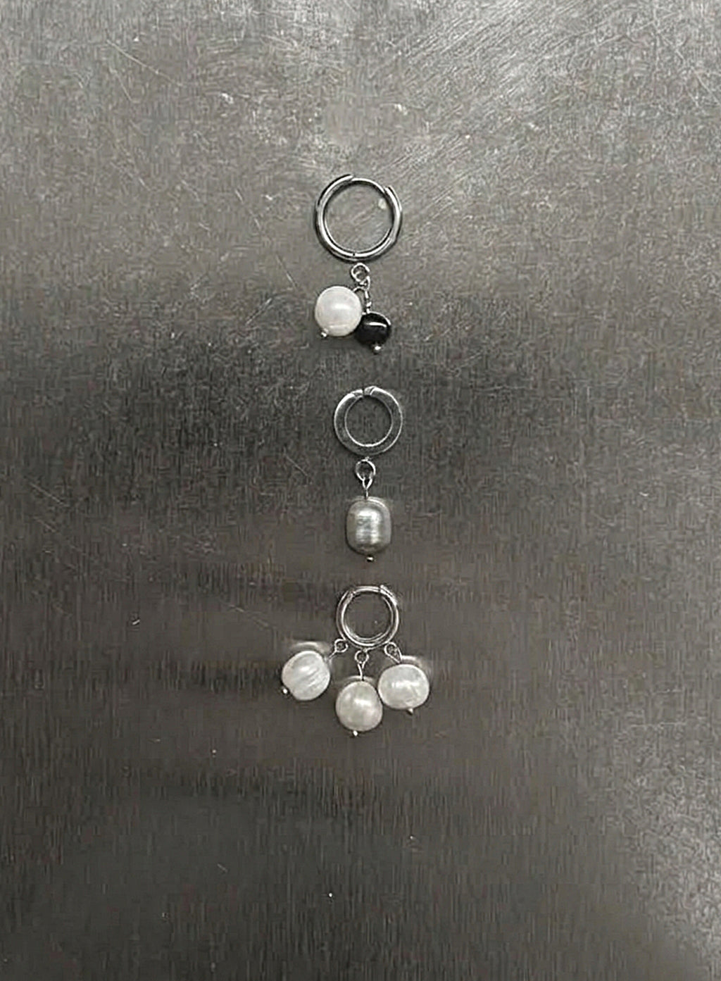 Three silver-toned ear cuffs with dangling pearl-like beads and a small black bead, arranged vertically on a dark gray textured surface.
