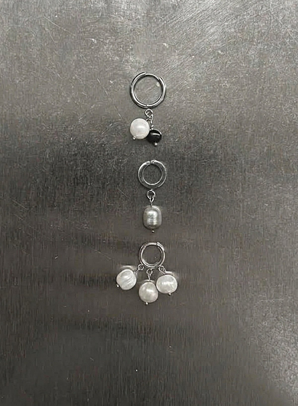 Three silver-toned ear cuffs with dangling pearl-like beads and a small black bead, arranged vertically on a dark gray textured surface.