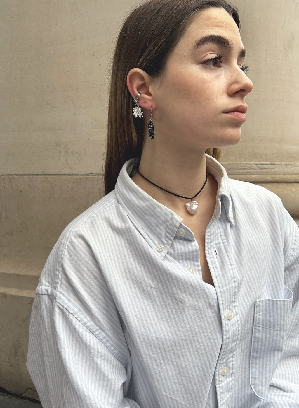 woman in stripe blue shirt wearing a cord necklace with a baroque pearl charm, multiple stone earrings against a beige stone wall