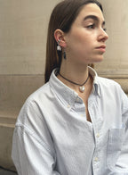woman in stripe blue shirt wearing a cord necklace with a baroque pearl charm, multiple stone earrings against a beige stone wall