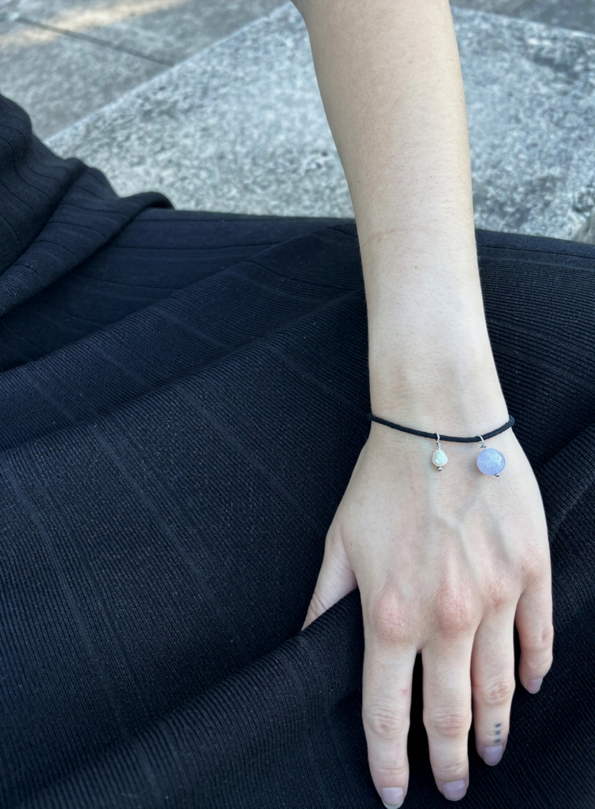 woman in black dress wearing black cord bracelet with blue stone charm and pearl charm against concrete background