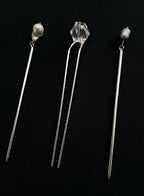 Three long silver hair pins displayed on a black background. The left and right pins feature small white pearl beads at the top, while the center pin has a faceted clear crystal bead.