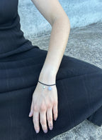 a woman in black dress seated and wearing black cord bracelet with blue stone charm and pearl charm against concrete background