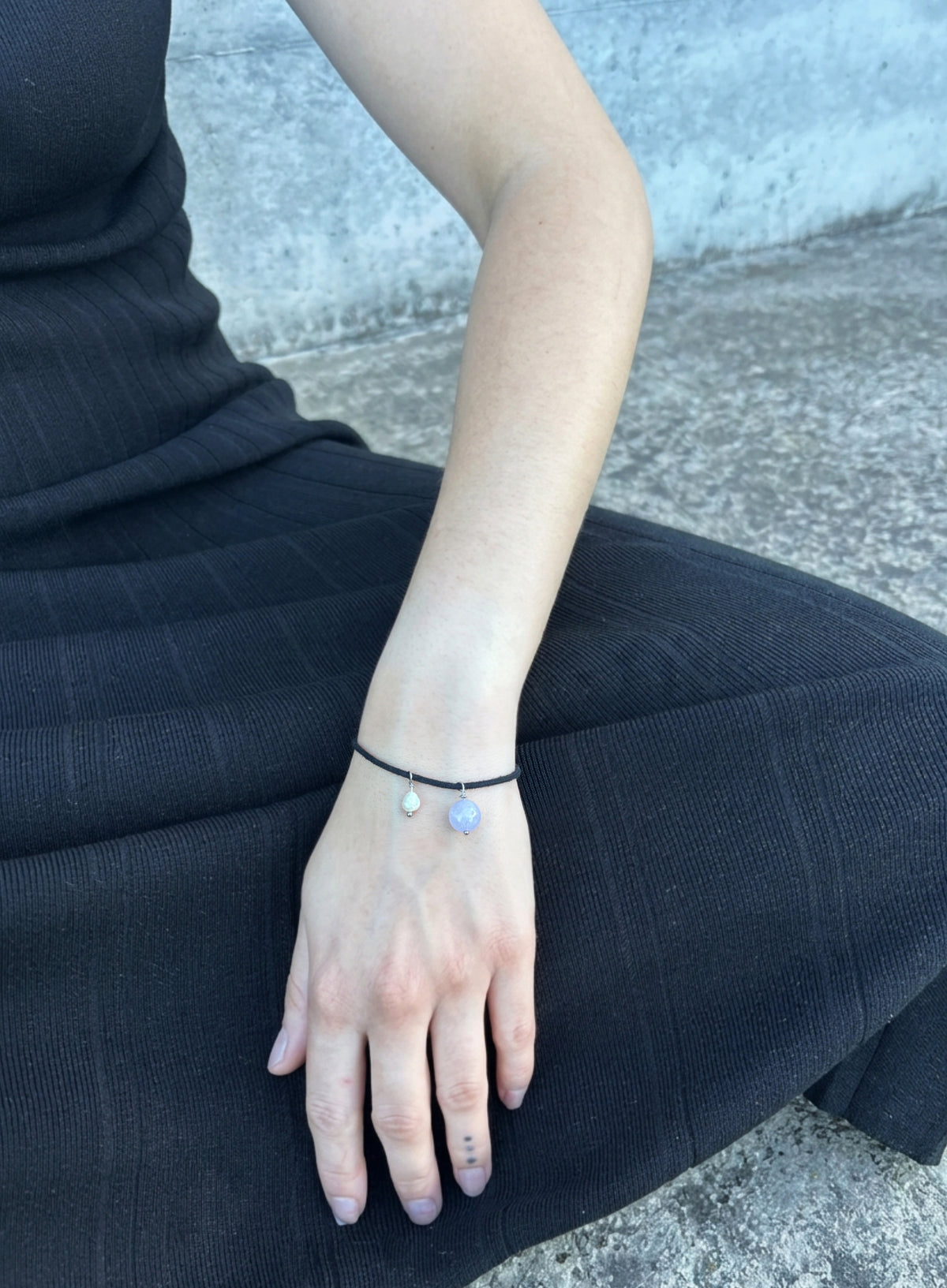 a woman in black dress seated and wearing black cord bracelet with blue stone charm and pearl charm against concrete background