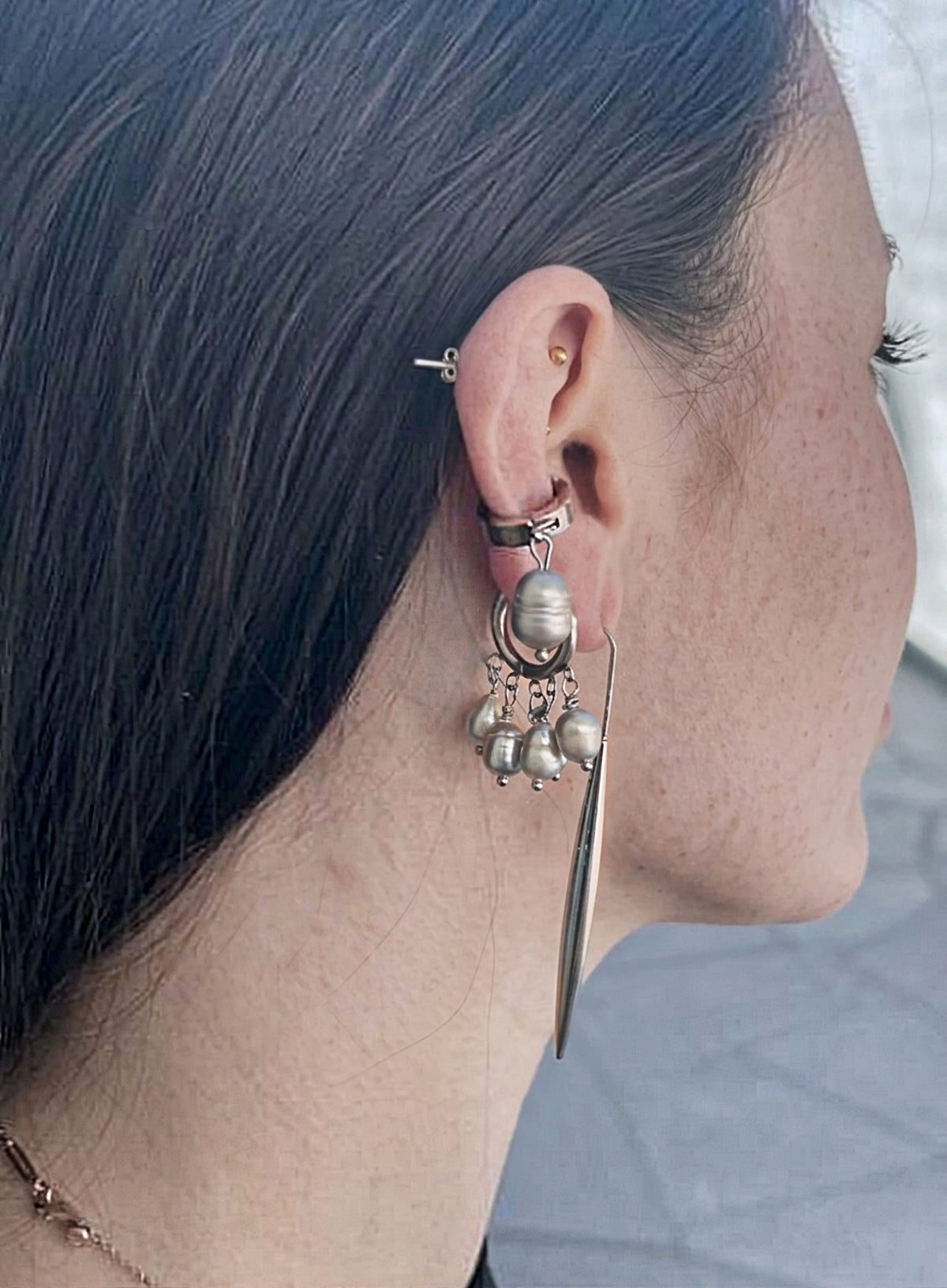 woman wearing a pearl of grey pearl charms earrings in huggie hoop, long structural shape earring, and a large grey pearl ear cuff against concrete background