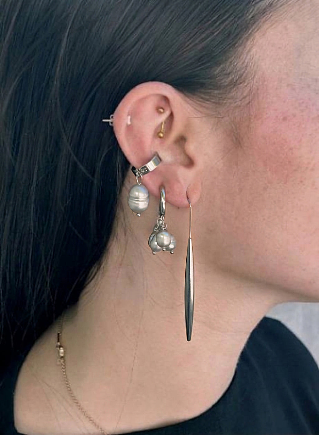 close up of a woman wearing a pearl of grey pearl charms earrings in huggie hoop, long structural shape earring, and a large grey pearl ear cuff against concrete background