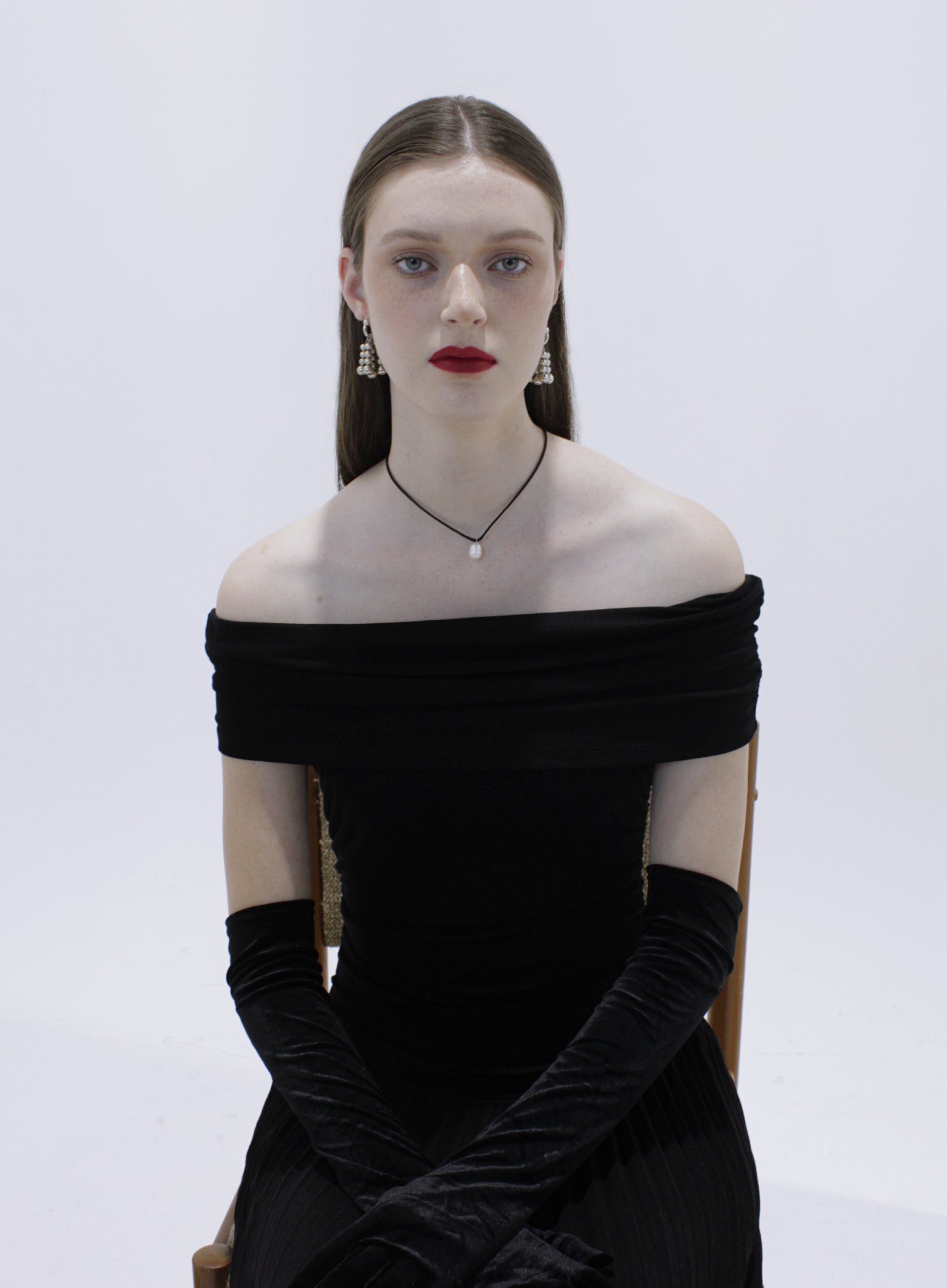 Woman wearing a black off-shoulder dress with long gloves and silver earrings with pearl chocker on a white background