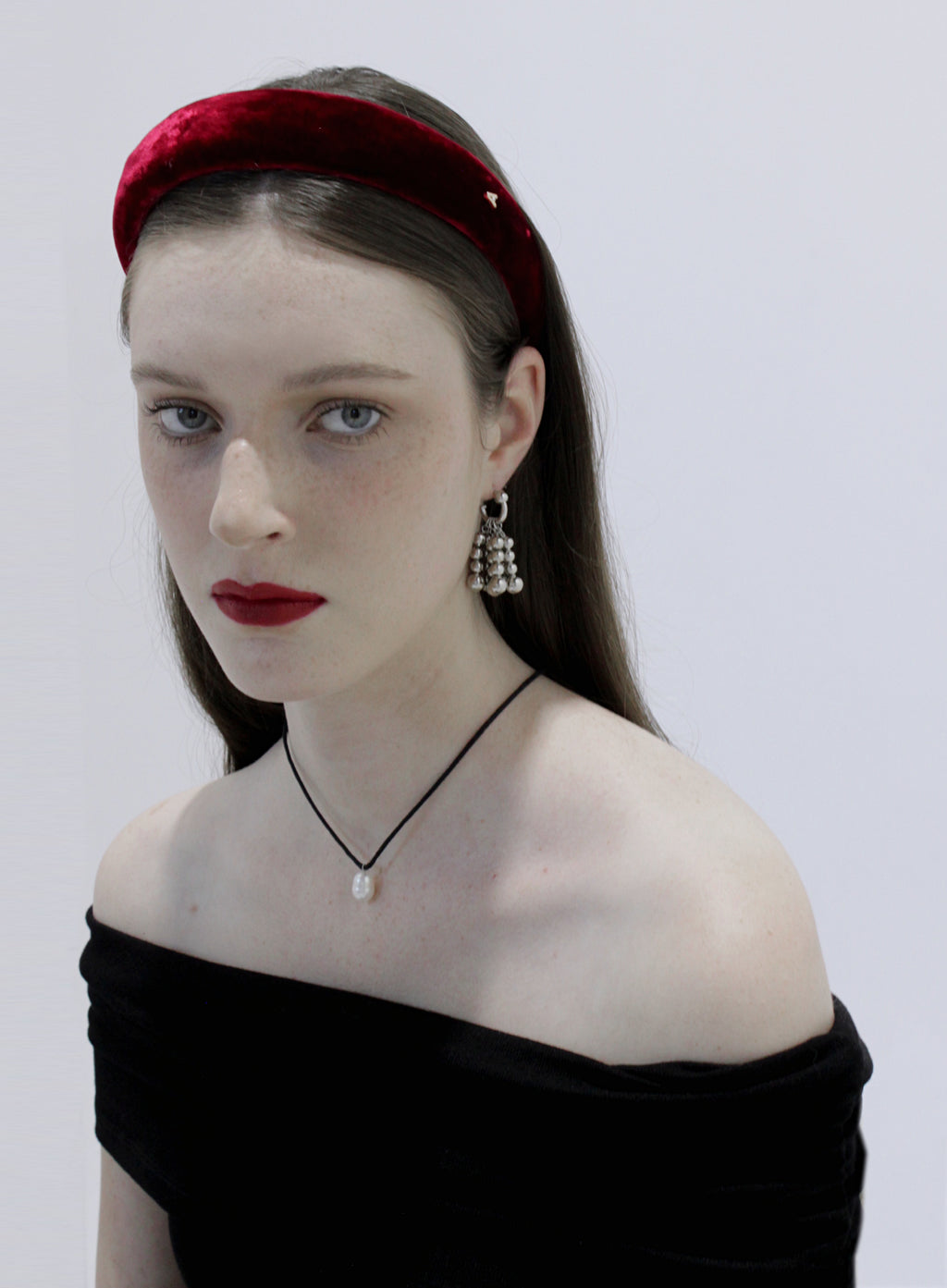 Woman wearing a red velvet headband, black off-shoulder top, and dangling earrings and black chocker with pearl pendant