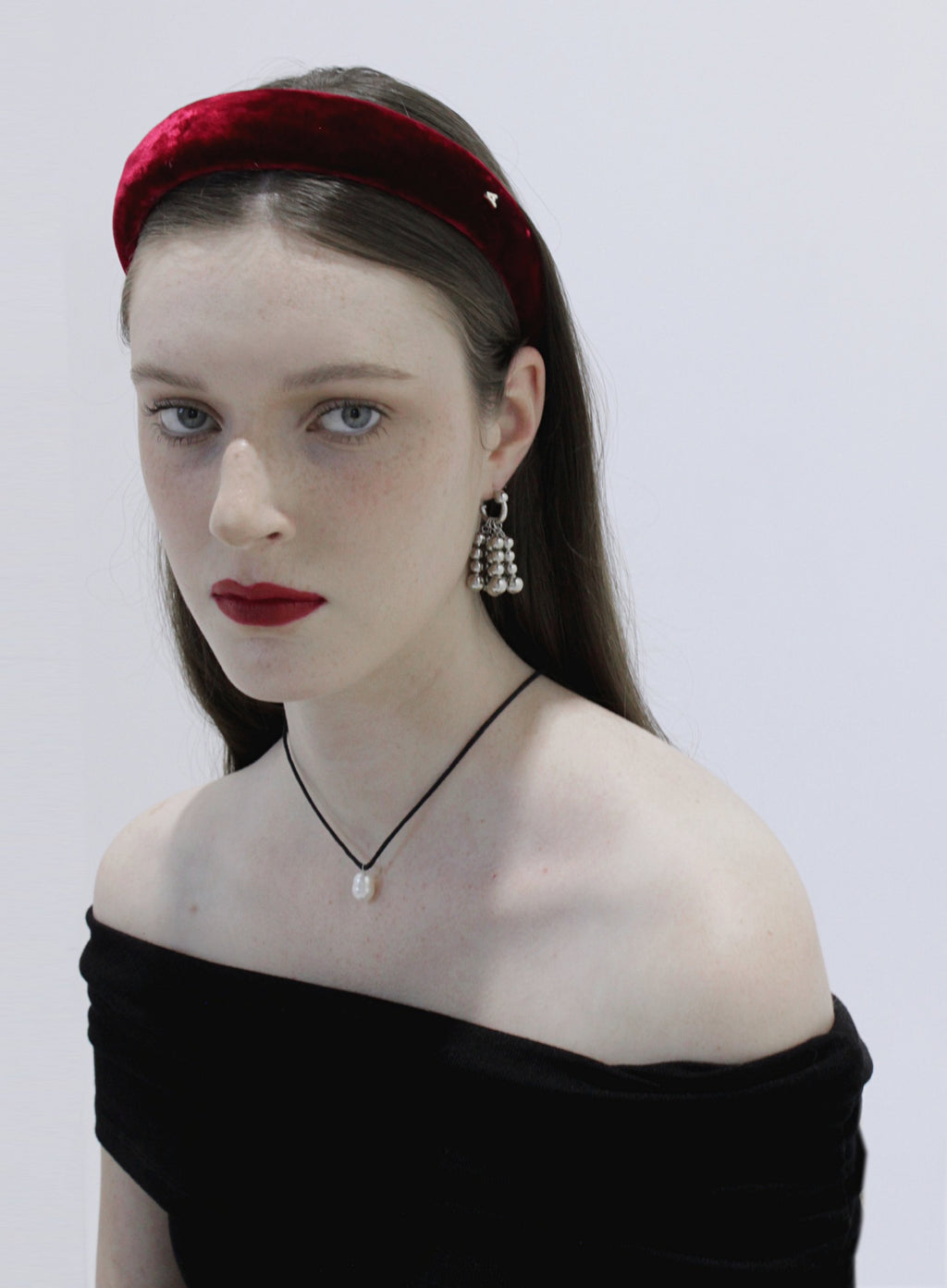 Woman wearing a red velvet headband, black off-shoulder top, and dangling earrings against a white background.
