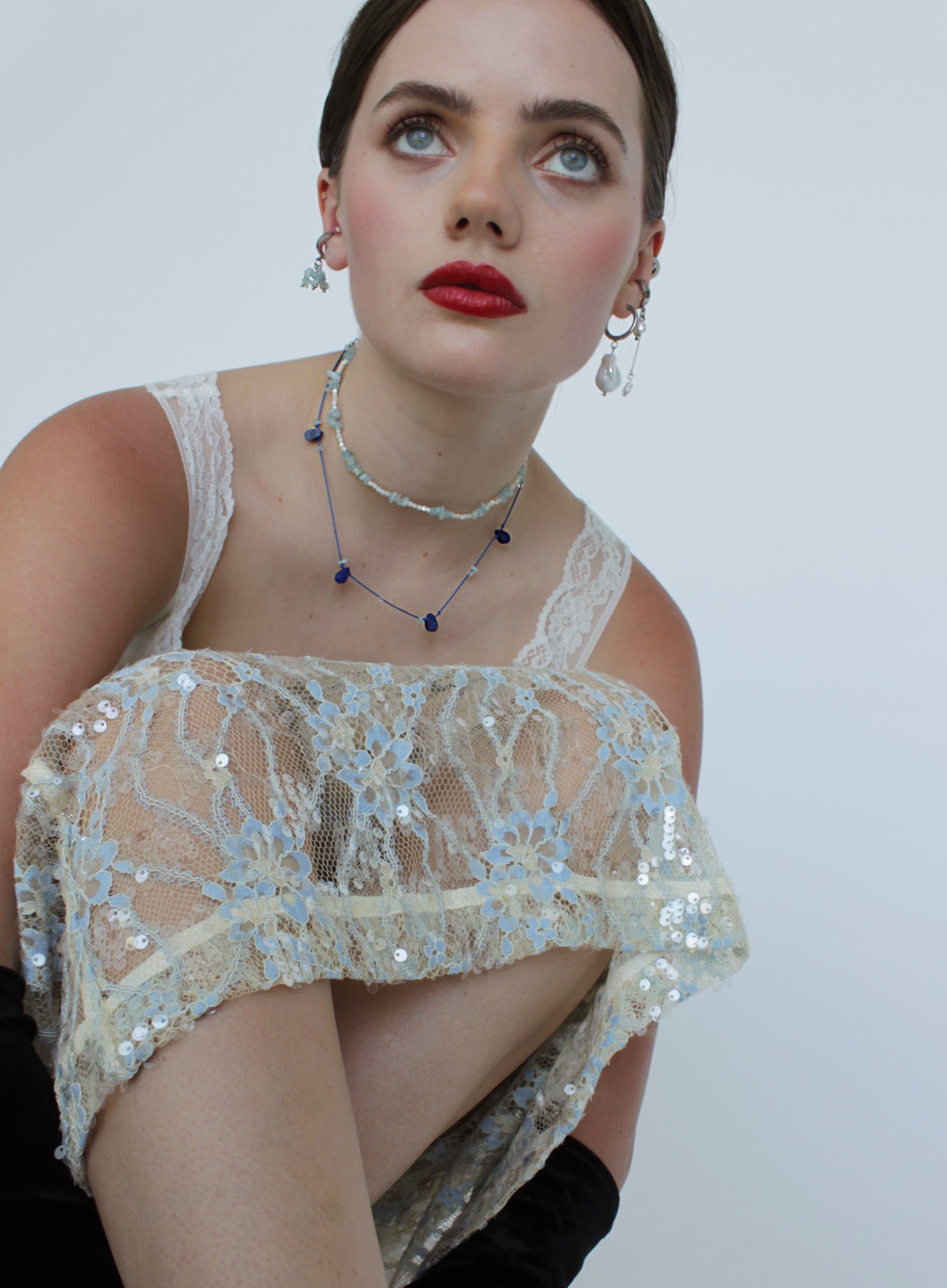 A close-up of a model wearing a sheer lace and sequin off-the-shoulder top in cream and light blue, paired with blue beaded necklaces and silver earrings, against a plain light gray background.