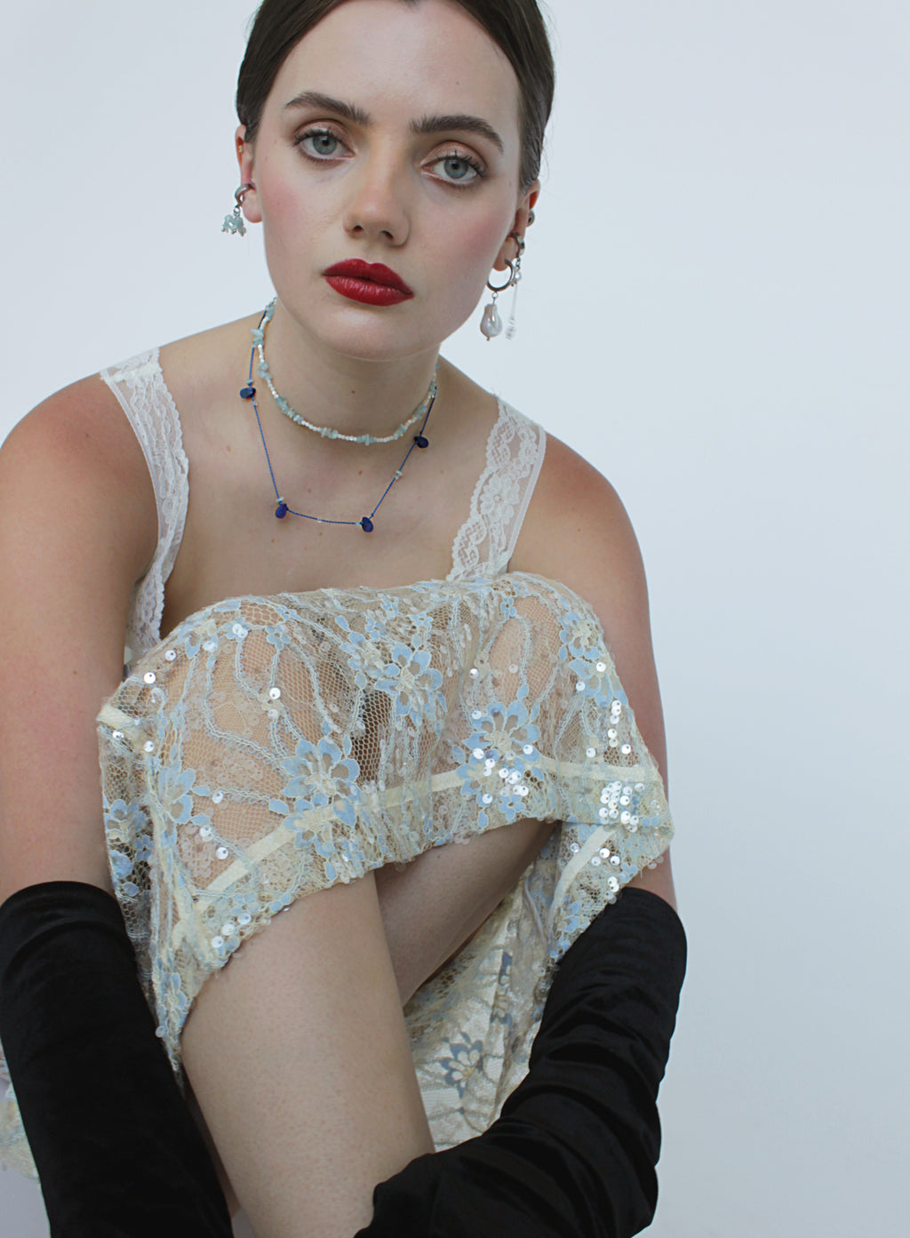 A model wearing a cream lace and sequin strapless top with black long gloves, layered necklaces, and dangling earrings, posed against a plain light gray background.