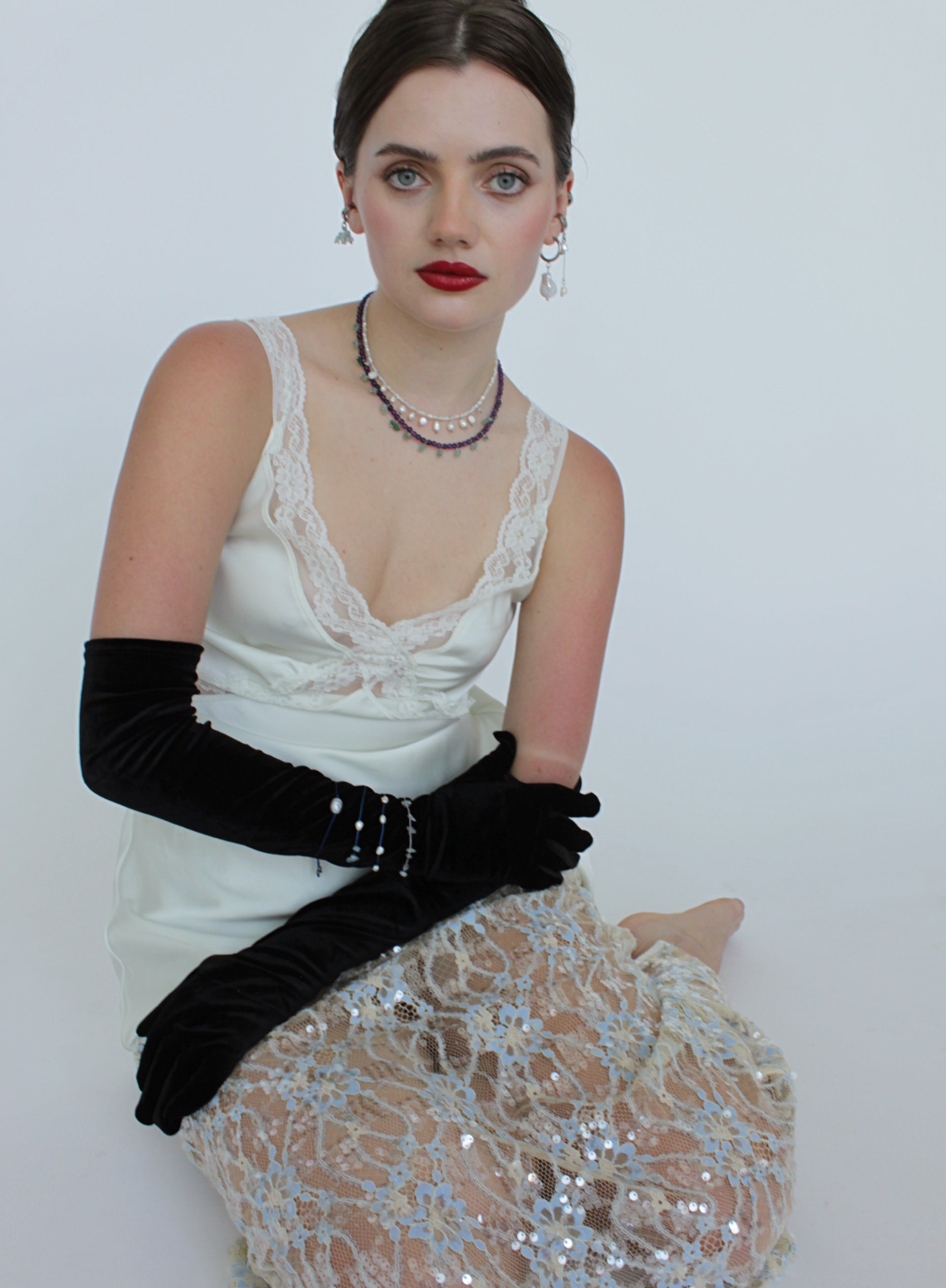 Woman wearing doubled layered necklaces on white dress with a deep neckline, black gloves, and a sequined skirt on a plain background