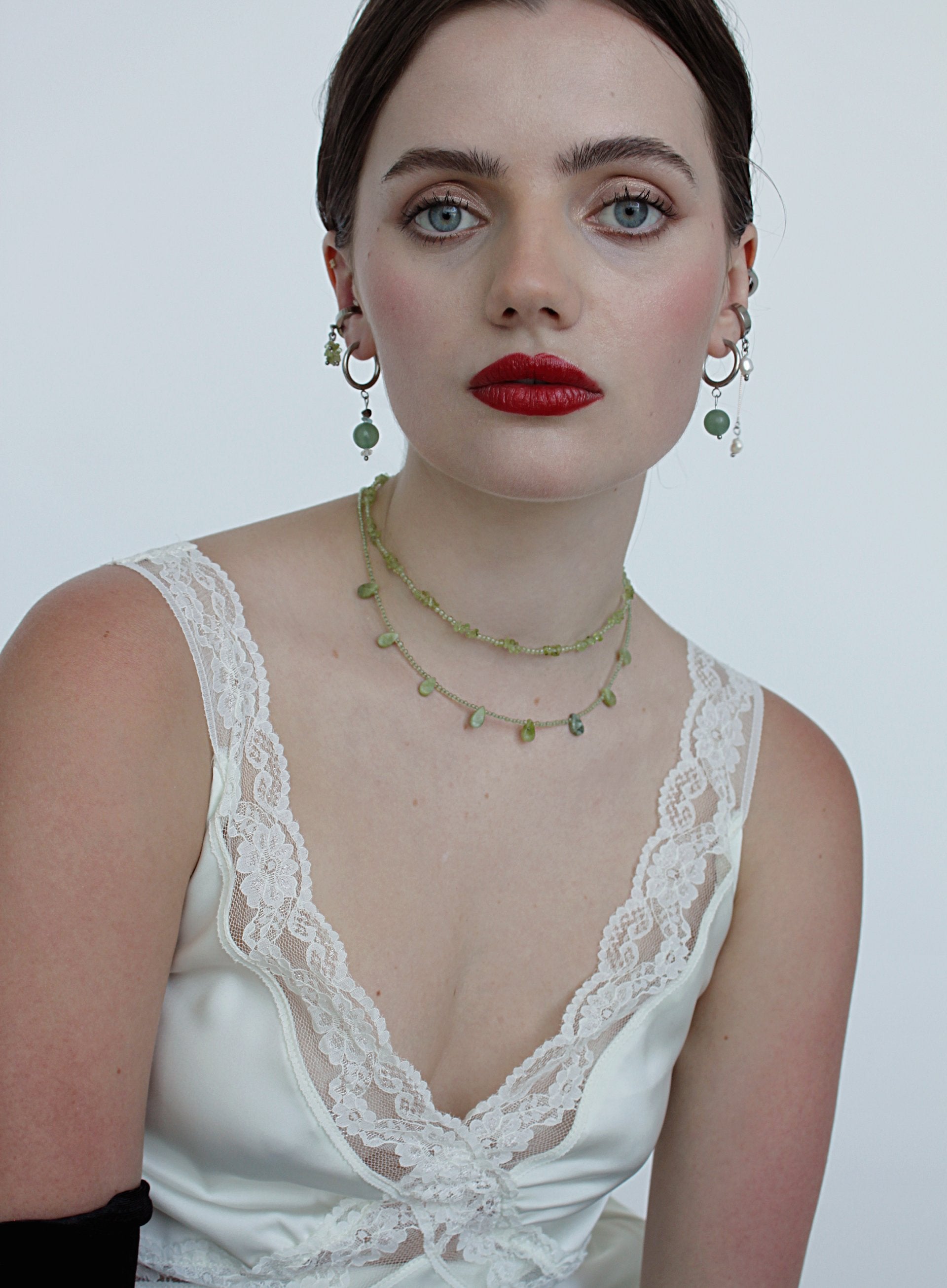 Woman wearing a white lace top with green jewelry against a plain background