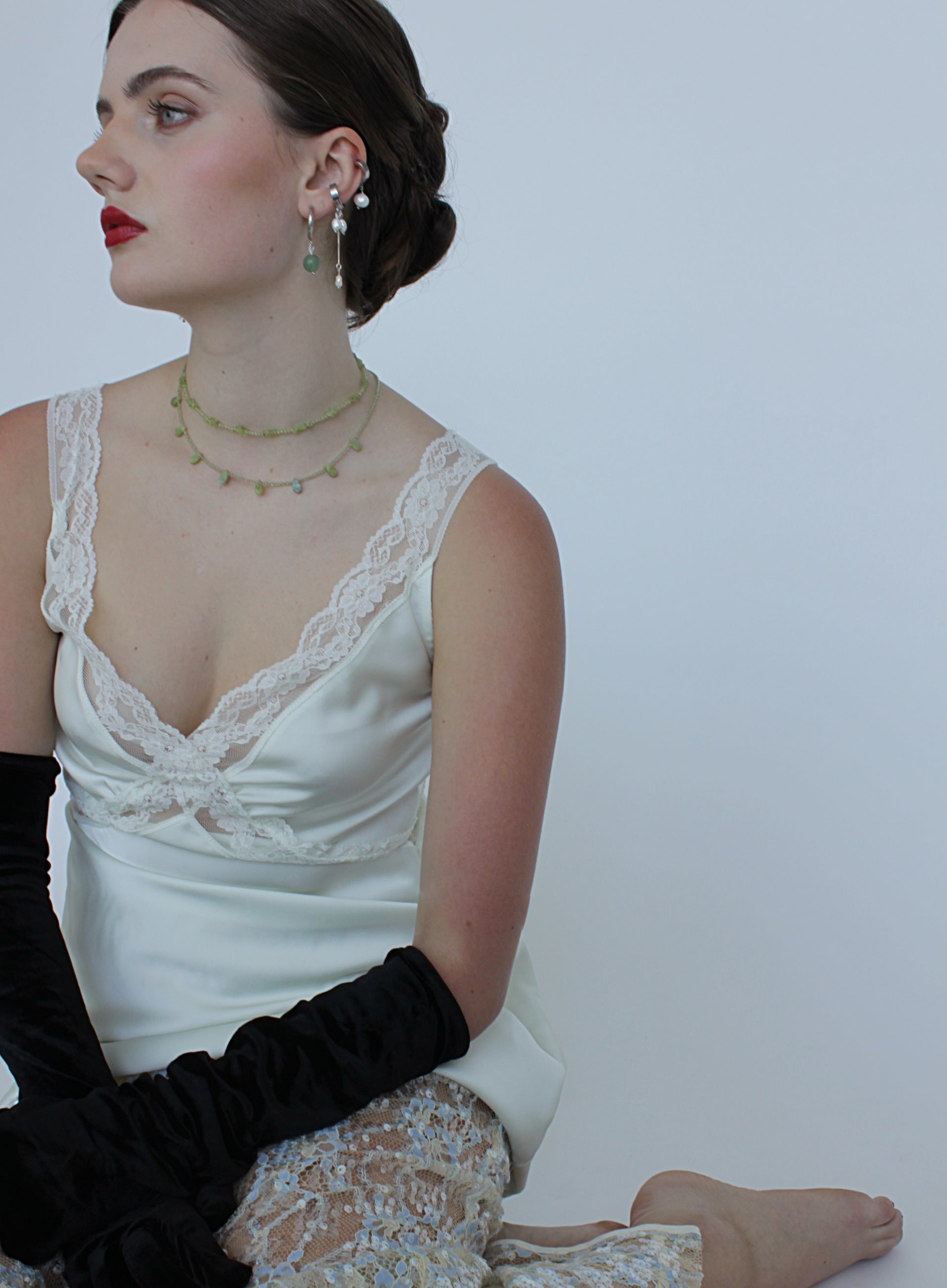 Woman wearing a white lace top, green necklaces and black gloves against a plain background