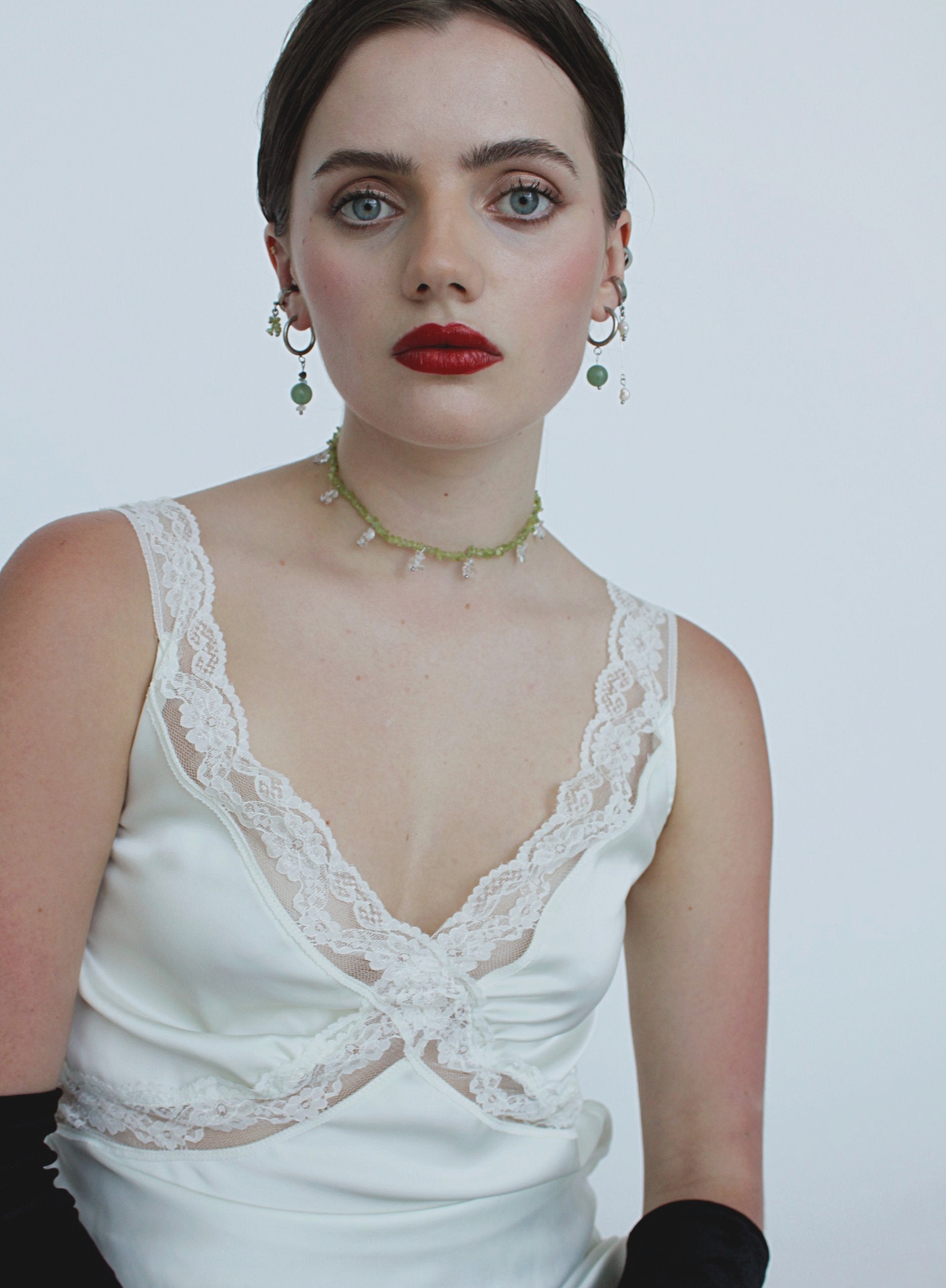 Woman wearing a white lace top with and green necklace on a plain background