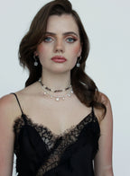 A woman with wavy brown hair and blue eyes wearing a black lace camisole top and layered pearl/beaded necklaces, photographed against a plain light gray background.