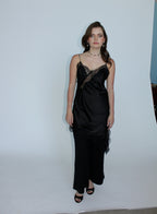 A woman modeling a black satin slip dress with lace trim and a draped, gathered skirt, standing against a plain white studio backdrop.