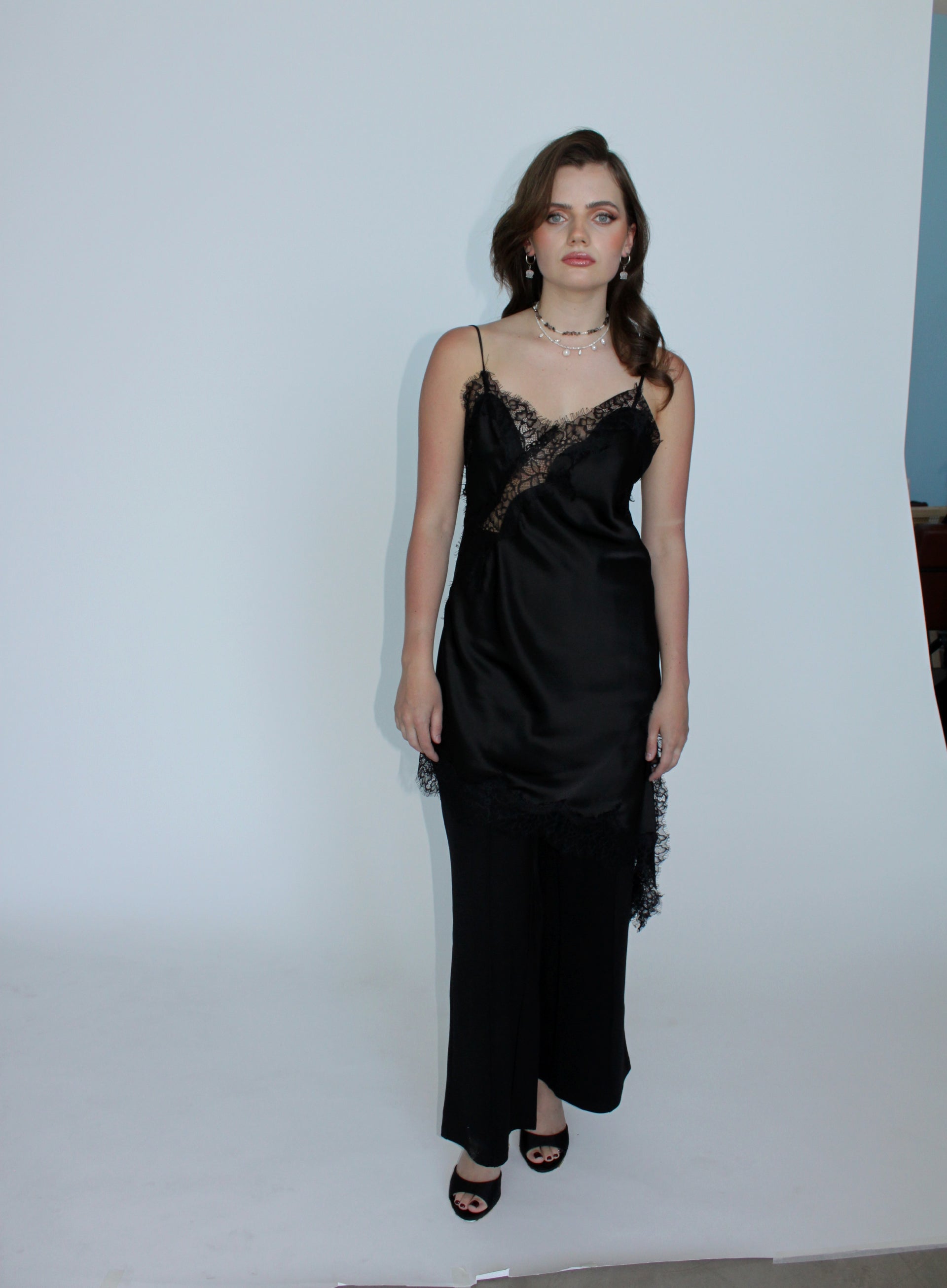 A woman modeling a black satin slip dress with lace trim and a draped, gathered skirt, standing against a plain white studio backdrop.