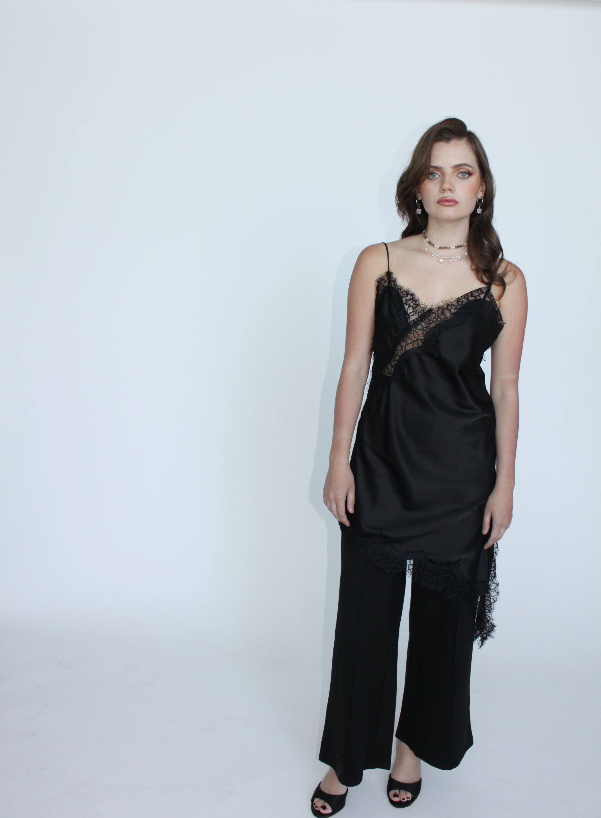 A woman modeling a black satin camisole top with lace trim and matching wide-leg pants against a plain white studio background.