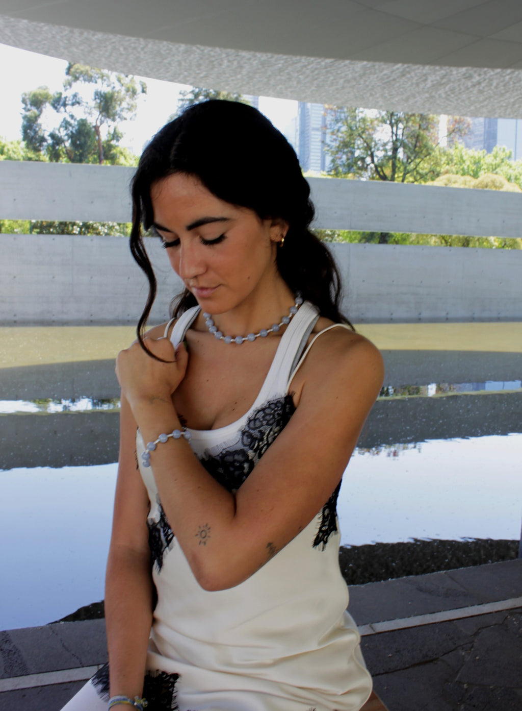 Woman sitting on a bench by a waterfront with trees and buildings in the background with blue stones bracelet and necklace
